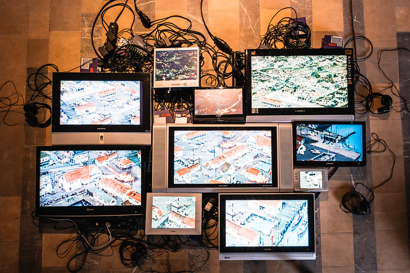 Several screens are assembled into an art installation, showing images of buildings and piers, with tangled wires visible in the background.
