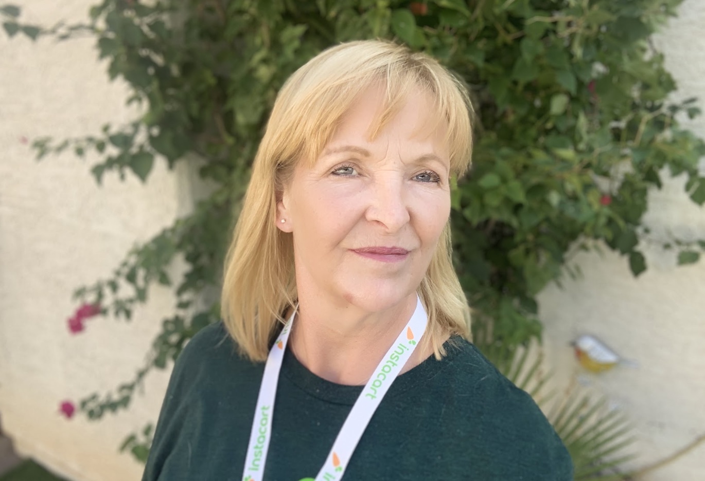 Photo of a blonde older and wiser woman wearing an Instacart lanyard in front of a wall with ivy